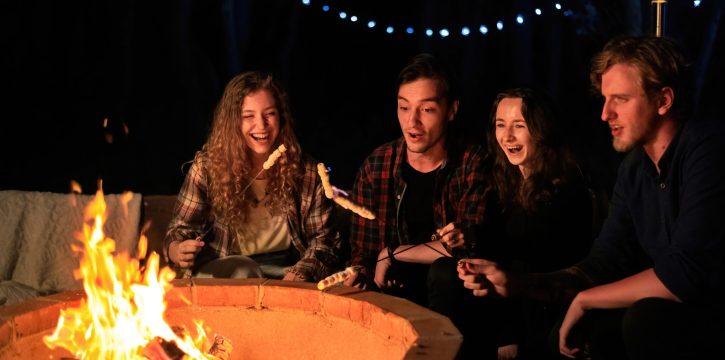 A group of happy young friends near a campfire at glamping, night. Two men and women. Frying marshmallow